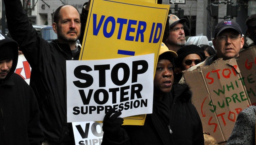 Photo showing people on a demonstration holding placards that say 'Stop Voter Suppression' and other messages which are partially obscured.