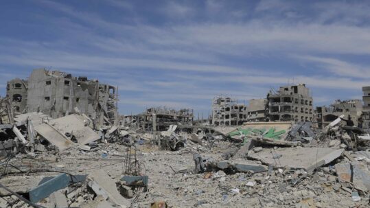 Photograph showing rubble in the foreground and around five destroyed multi-storey buildings in the background. The sky is blue with some whispy clouds.