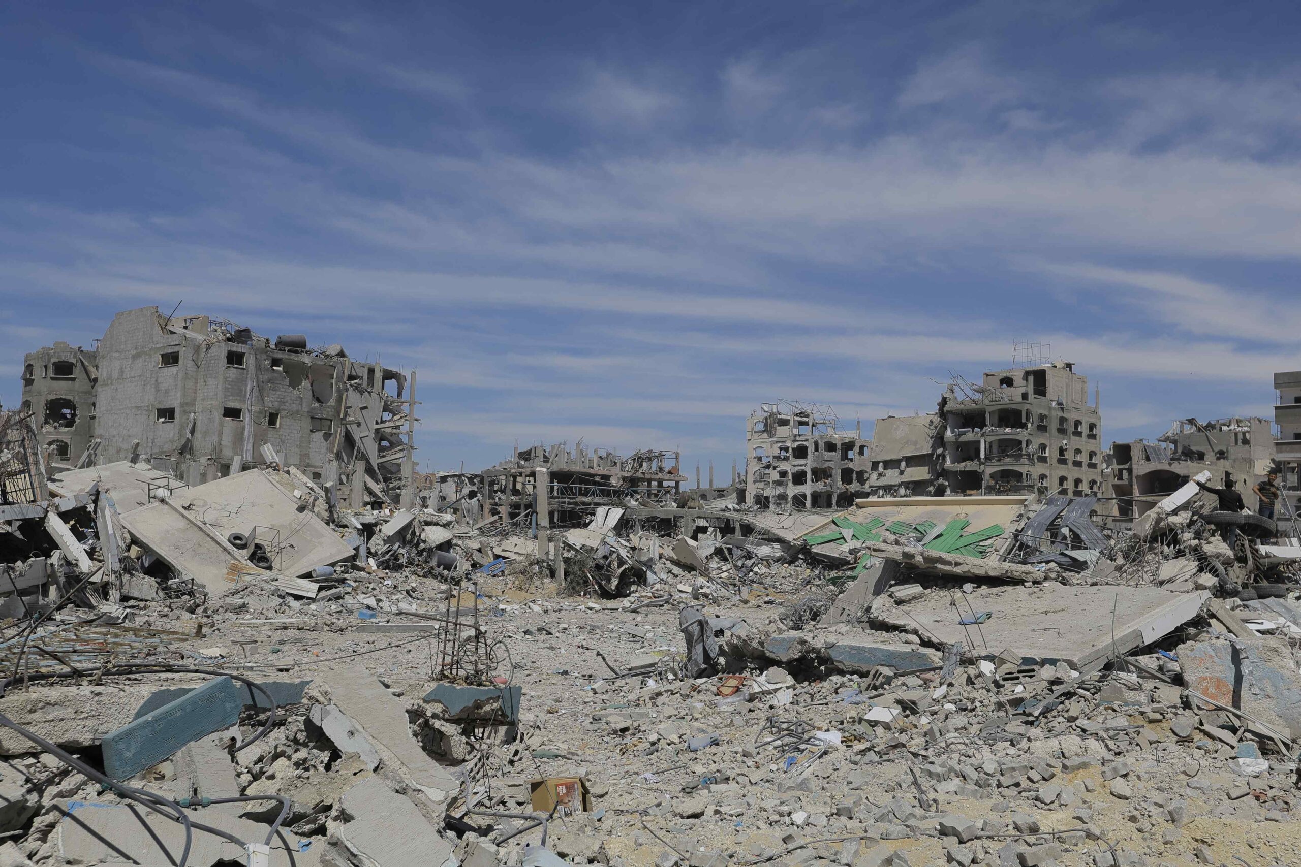 Photograph showing rubble in the foreground and around five destroyed multi-storey buildings in the background. The sky is blue with some whispy clouds.