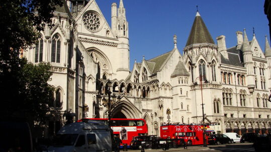 Photograph shows the front of the Royal Courts of Justice with two red buses in the foreground, against a blue sky.