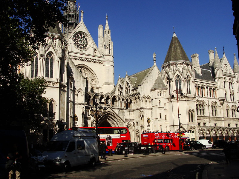 Photograph shows the front of the Royal Courts of Justice with two red buses in the foreground, against a blue sky.